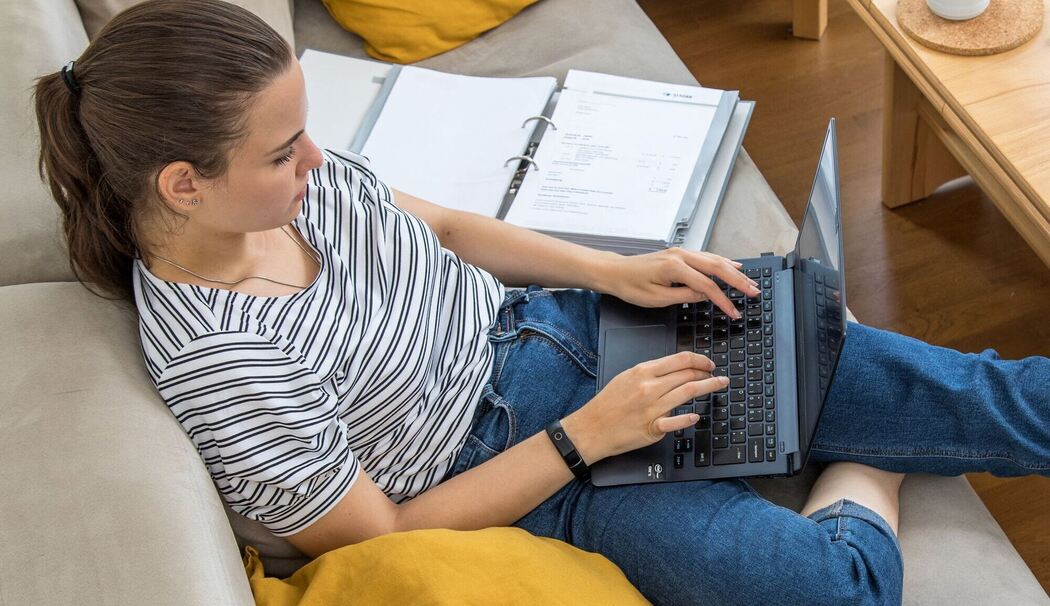 Eine Frau sitzt mit Laptop und Aktenordnern auf einem Sofa