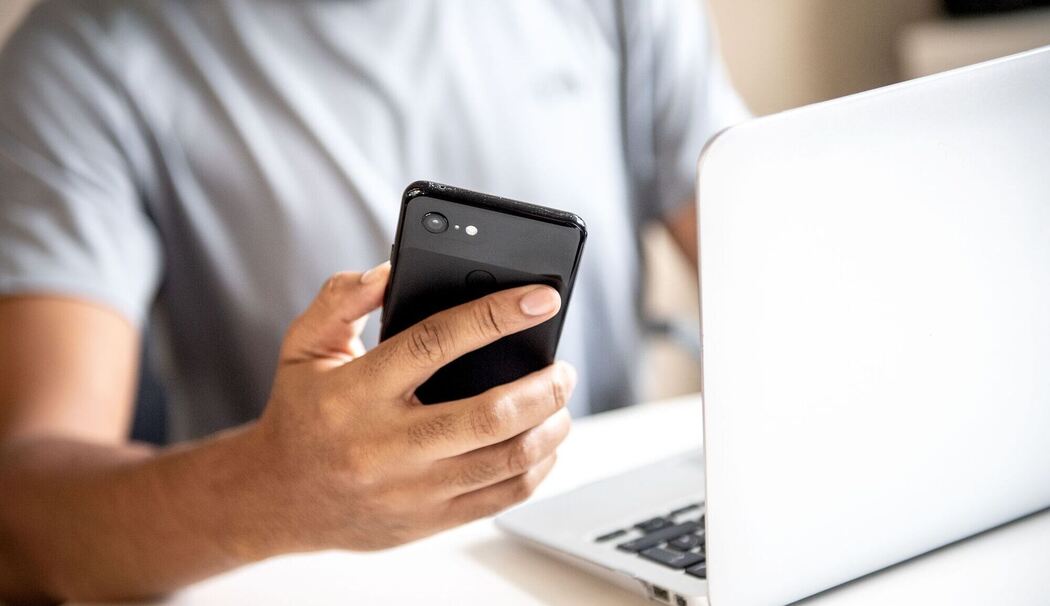 Mann hält vor einem Laptop sein Smartphone in der Hand